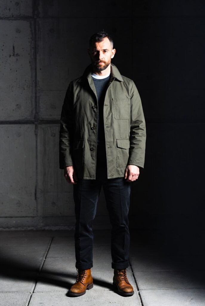 Man in charcoal merino sweater, olive chore coat, and brown boots showing comfort that serves presence — structured and sharp