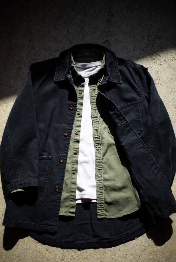 Layered navy chore coat over olive shirt and white tee on concrete — structure through layering