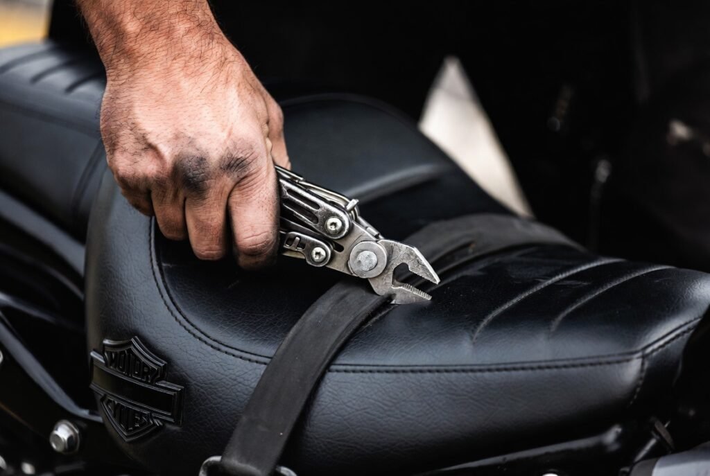 Close-up of a man’s callused hand using a well-worn multi-tool to tighten a tank strap on a Harley-Davidson style motorcycle seat. Natural light, gritty masculine maintenance shot. Tools that solve problems, not collect dust.