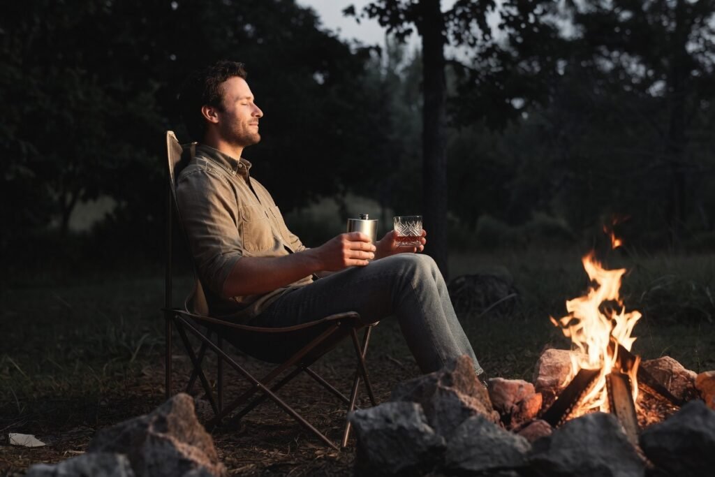 Man in quality button-up shirt and straight leg jeans sitting in a folding camp chair to the left of a fire pit in a woodsy setting. Subtle content expression, holding flask and vintage whiskey glass. Dark moody tones, minimalist composition focused on joyful solo competence. No logos — pure signal.