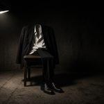 The Performative Man Empty dark suit on a wooden chair in a hard-lit room, no wearer, concrete floor.